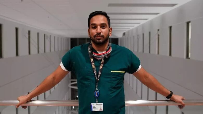 A man stands in green scrubs against a railing. 