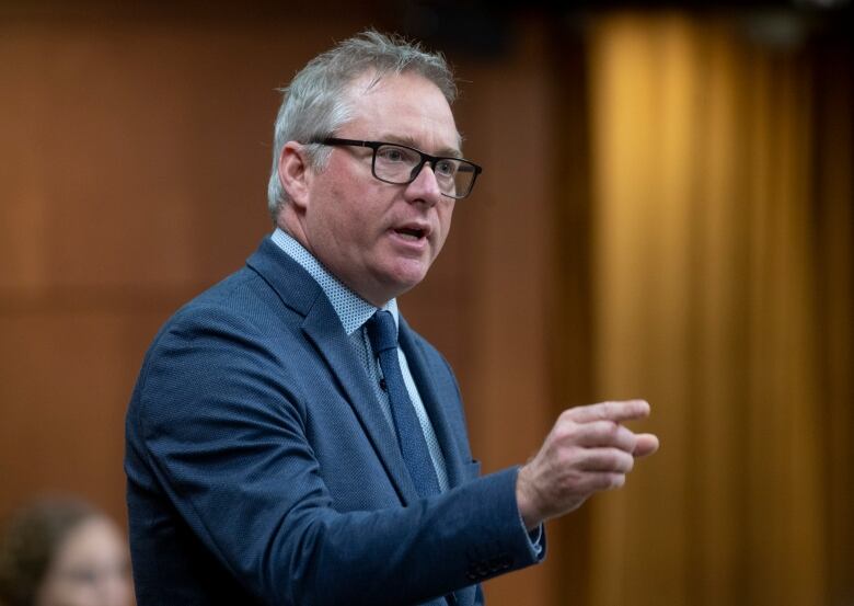 A man in glasses and a blue suit stands and speaks.