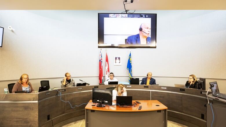 Politicians and administrators sit behind a curved council table in a meeting room.