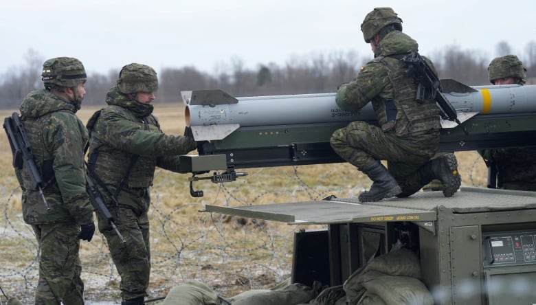 Four men in green camouflage military wear and helmets stand near a large missile, which is parallel to the ground and sitting on a large piece of green machinery. The soldiers, who have guns slung onto their backs, stand near razor wire in a faded green field. A treeline is visible in the distance.