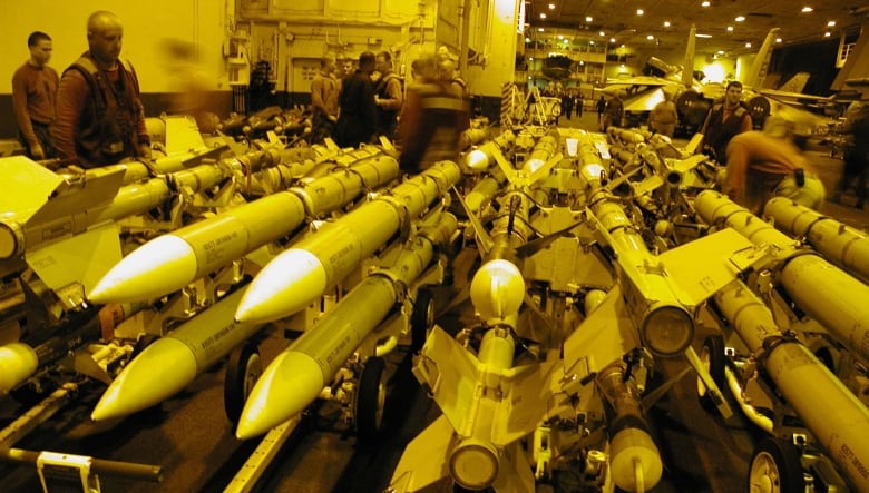 In a photo with a yellow tint, people stand near scores of long, slender missiles with stabilization fins visible on some. A fighter jet is visible in the background.