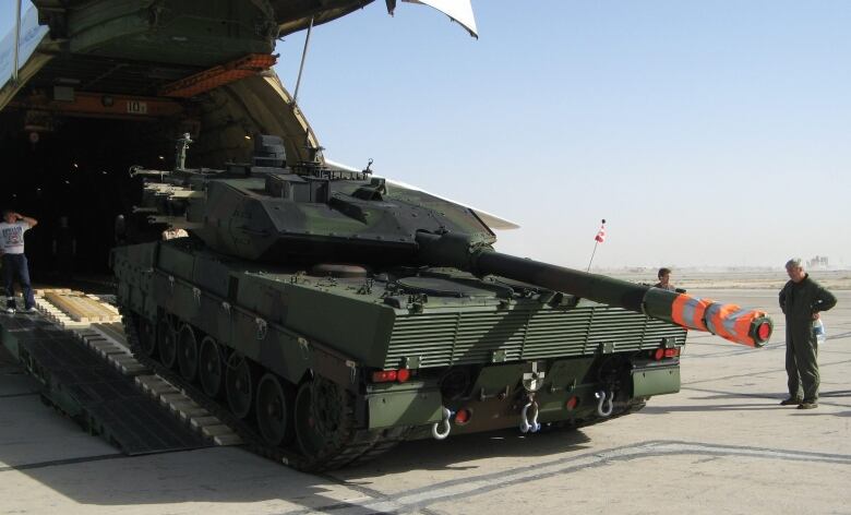 A large green tank rolls off a ramp from an airplane as three people look on.