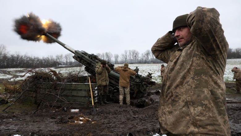 Five men in military camouflage hold their hands over their ears as a large gun fires a round. Smoke and flames are visible exploding from the barrel. The scene is a muddy area near a field covered in light snow.