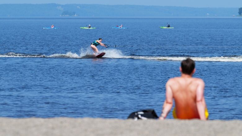 A man sits on a beach while a woman water skis and people paddle kayaks on a river.