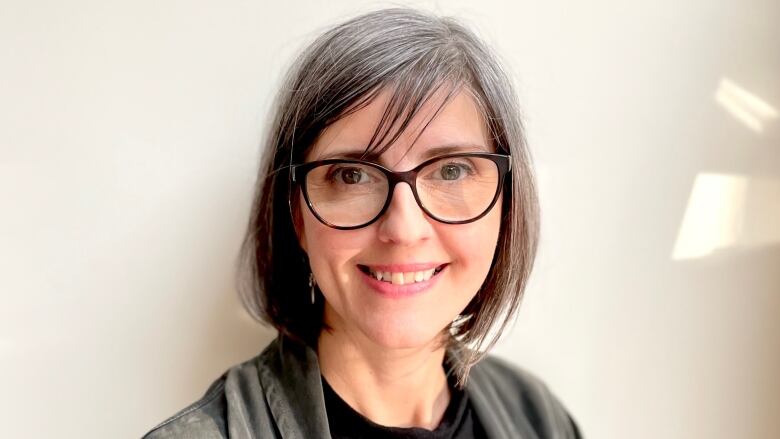 A woman with straight hair down to her chin-level is smiling and wearing glasses. She is wearing a mock-neck top with an open cardigan on top.