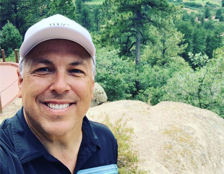 A man in a blue shirt and baseball cap stands by a rock at the top of a mountain with trees in the background.