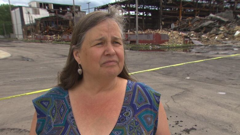 A woman looks forward, standing in front of a large warehouse destroyed by a fire.