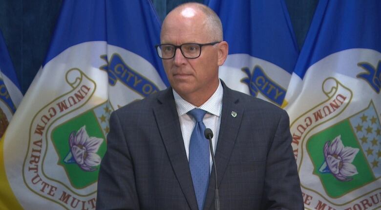 A man wearing black rimmed glasses and a navy suit stands in front of two city of Winnipeg flags.