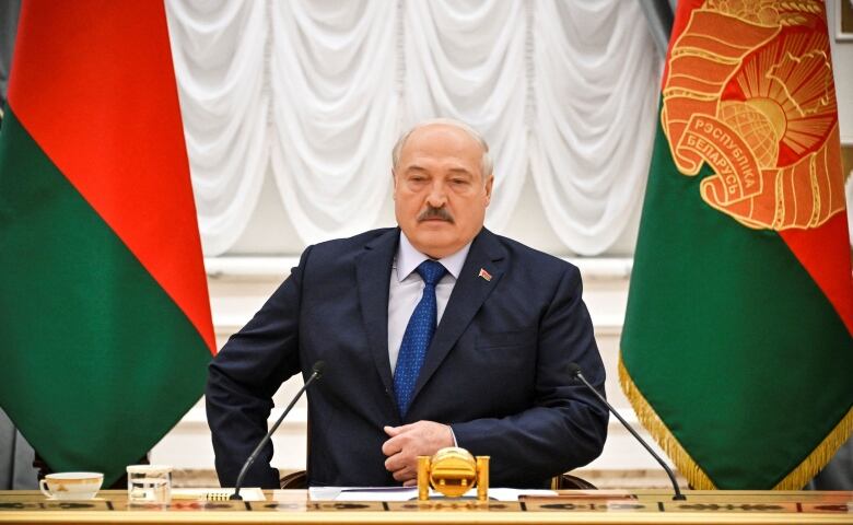 A man in a suit is shown seated at a table, with giant national flags behind him.