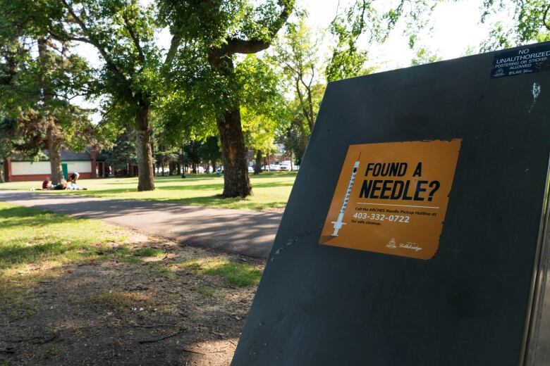 A green garbage receptacle in a tree lined park with an orange sticker reading 