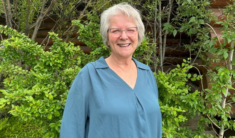 A woman with short grey hair and a blue long sleeve shirt standing in front of bushes.