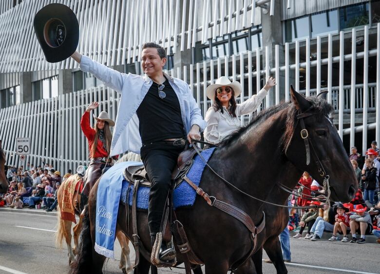 a man and a woman side by side on horses wave at people as they ride down a city street