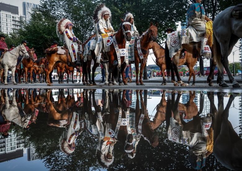 several people wearing traditional Indigenous costumes with headdresses sit on horses as their reflections show in a puddle in the foreground