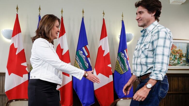 a woman on the left extends her hand with a man on the right for a handshake. Both wear western Stampede outfits. Behind them are Alberta and Canadian flags, six, alternating.