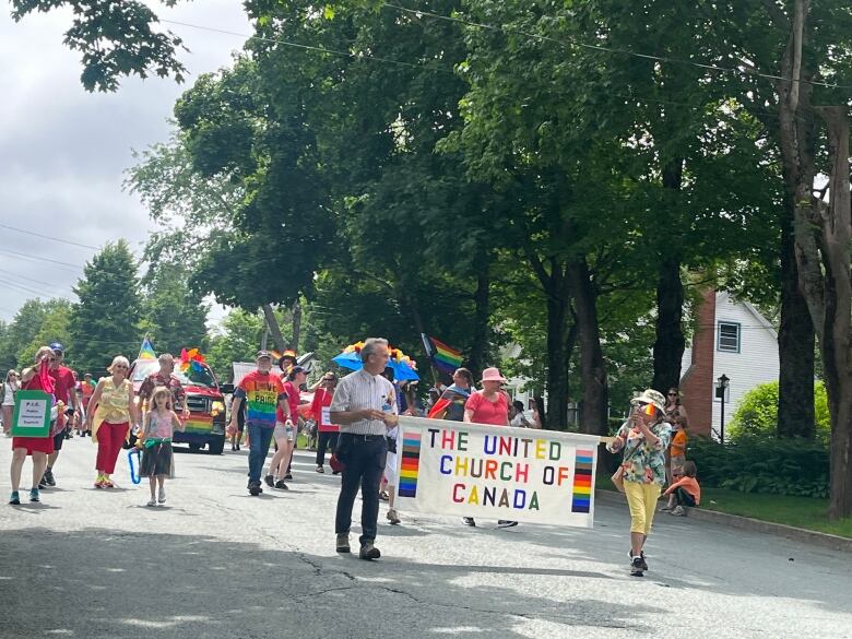 People with pride flags and colours walk on a street led by two people holding a banner saying United Church of Canada.