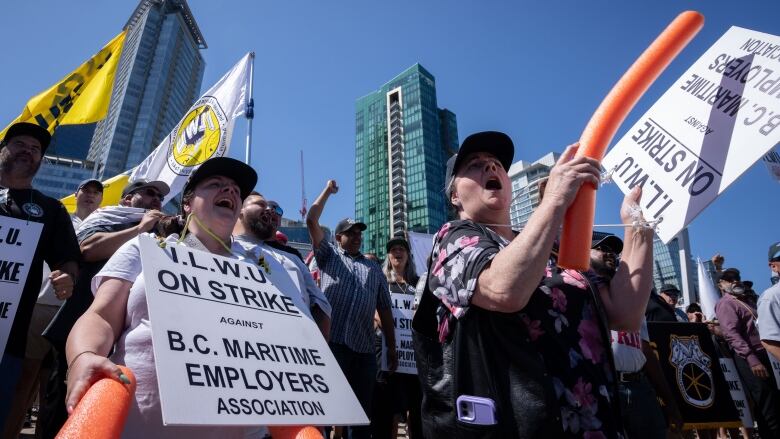 Workers wearing sandwich boards, carrying flags and holding balloons stand in a group in front of high-rise buildings in a city on a sunny day.