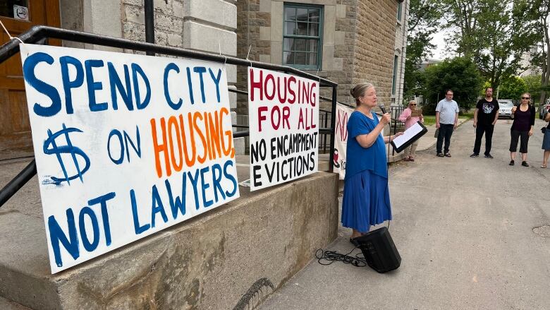 A woman in a blue dress speaks into a microphone. She's standing next to two signs, one of which says 