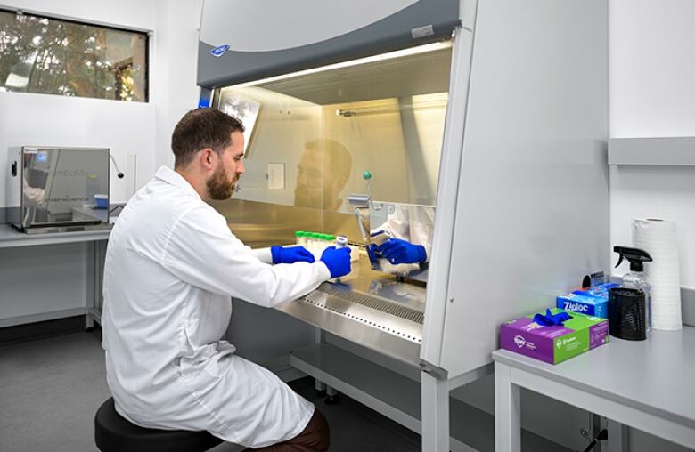 A white man with short brown hair and a short brown beard sits in front of a fume hood in a laboratory. He is wearing a white lab coat and blue gloves, and holding test tubes under the hood.