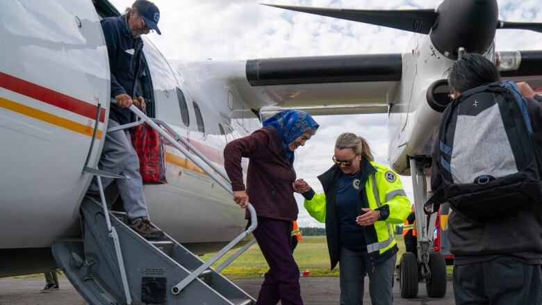 A younger woman in a safety vest helps and older woman off a plane. 