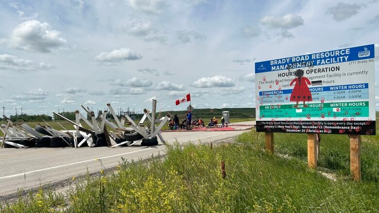 A sign with a person in a red dress painted on it is seen in the foreground. A blockade made of wood sticking out of tires is seen in the background on a road.