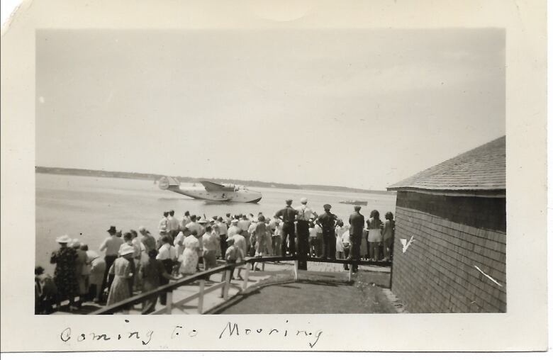 A black and white photo of a crowd of people on a wharf watching a large plane land on the water.