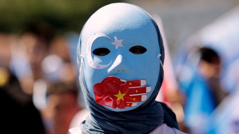 A person wears a blue mask over their entire face. The mask has a red hand drawn across the mouth and a moon and a star painted near the left eye. 