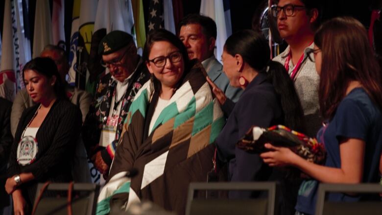 A woman is wrapped in a starblanket on a stage with other people.