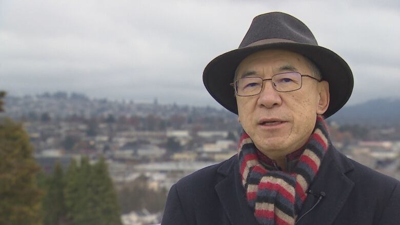 A man with hat stands with hills and buildings in the background