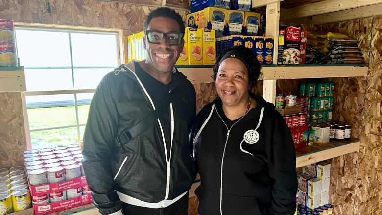 A man, on the left, and a woman, on the right, smile for a photo in front of shelves lined with non-perishable food.