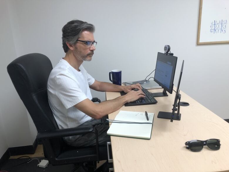 A man wearing a white t-shirt and grey pants sitting at his computer