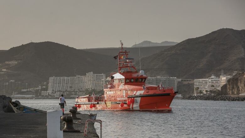 A red boat on the water sails past mountains and buildings on the coast. 