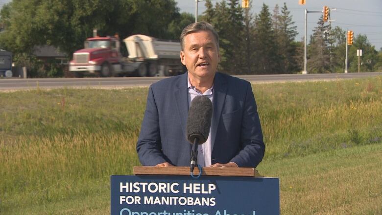 A man in a blue suit stands at a podium behind a microphone as a semi truck drives behind him. The podium has a sign that says, 