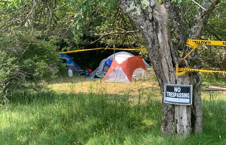 A 'no trespassing' sign hangs from a tree, with camping tents set up behind. Yellow police caution tape surrounds the scene.