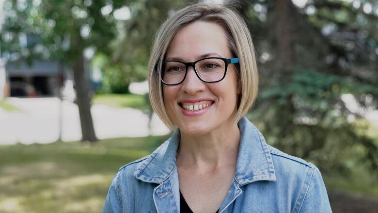 A close-up shot of a lady smiling. She's dressed in a denim shirt and is wearing thick black glasses.