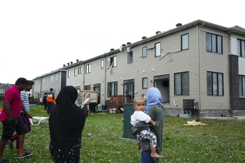 People stand outside a row of homes with hail and debris outside of it after a storm.