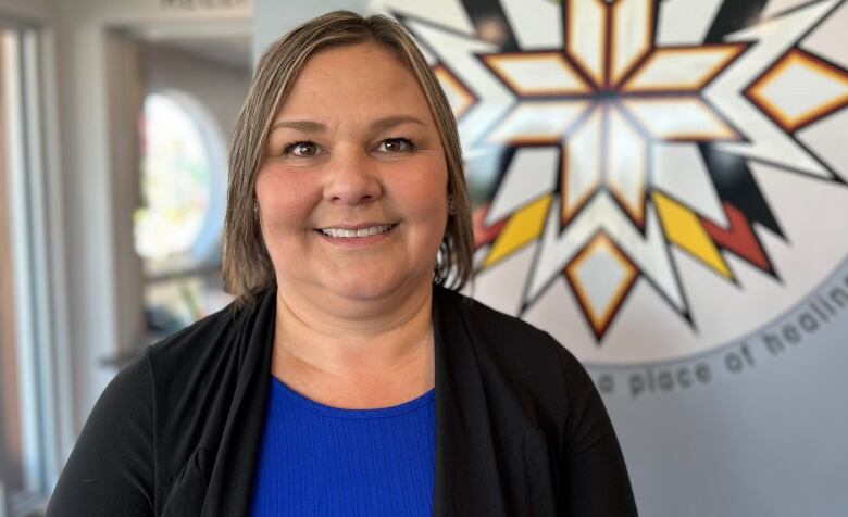 Shelly MacLean, a smiling woman with short blond hair, stands in front of a Mi'kmaw emblem painted on a wall. 