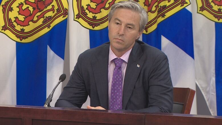 A man in a suit sitting at a table is backed by flags.