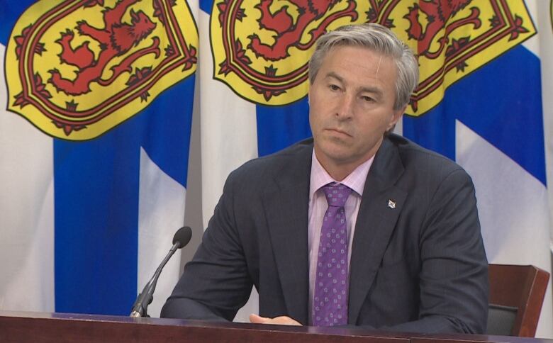 A man in a suit sitting at a table is backed by flags.