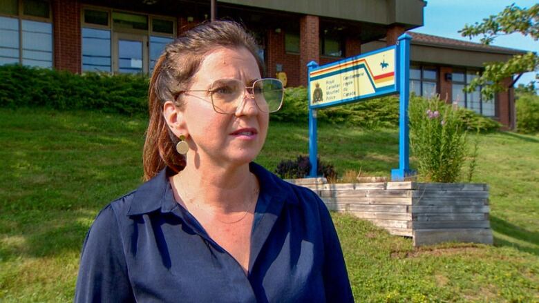 A woman with long brown hair tied back in a ponytail, wearing a blue shirt and sunglasses, standing in front of an RCMP detachment.