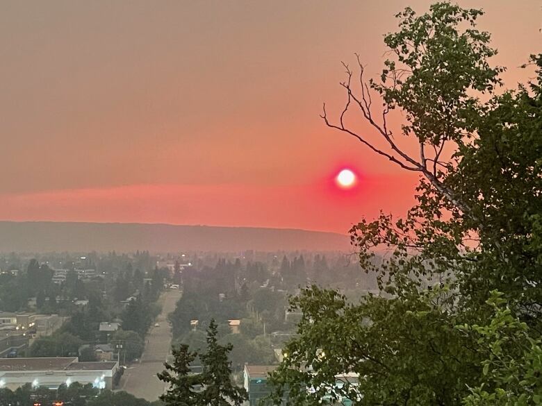 The sun is a pale orb seen through a blood-orange sky over a city and trees.