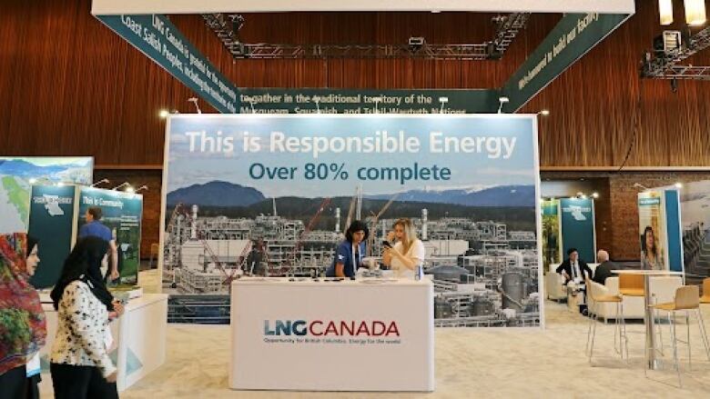 There is a booth for LNG Canada in a large room lined with wooden panels. Women dressed in blue and red/green hijabs are walking past. At the center of the booth is a woman in a blue shirt and a woman in a white shirt. A panel behind the women reads, 