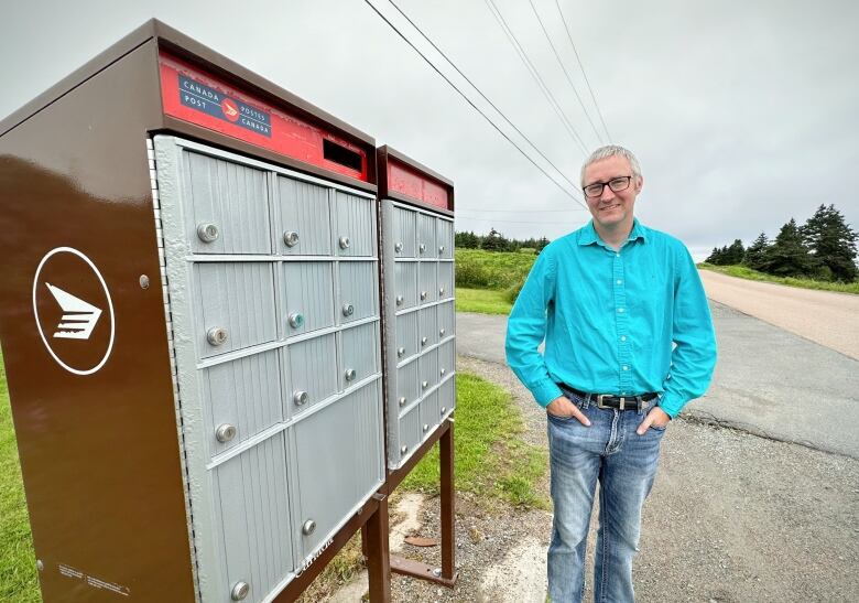 A man with a blue long-sleeved shirt and glasses stands next to a Canada Post community mailbox.