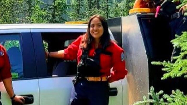 A smiling woman with dark shoulder-length hair wears a red button-up shirt and leans on the side of a white pickup truck in a forest.