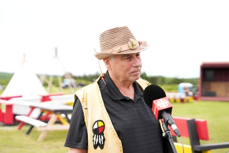 An older man wearing a fedora and a vest with Indigenous patterns stands in front of a microphone.