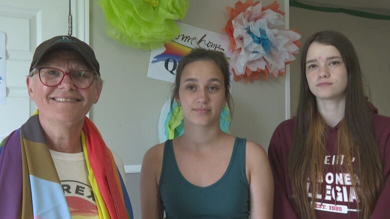 An older woman and two teenage girls are in front of pride themed rainbow decorations. 