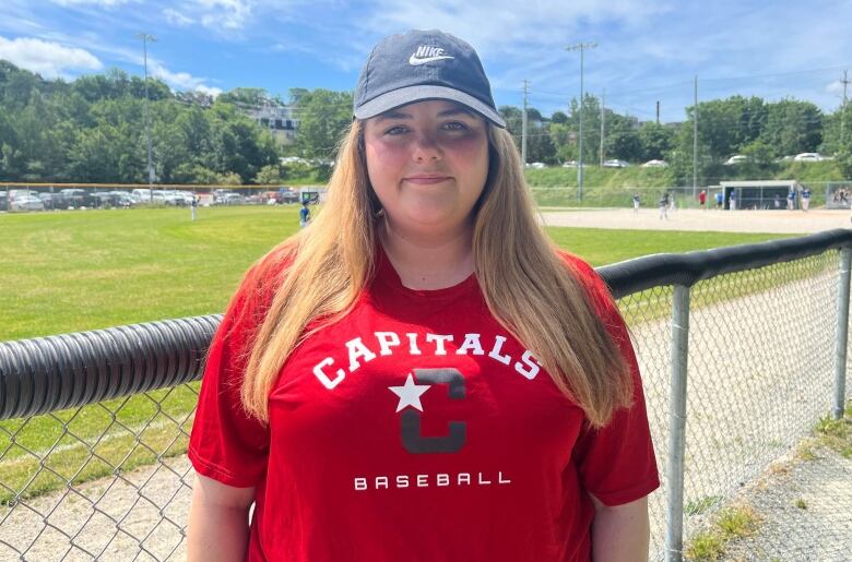 A blonde woman in a red Capitals Baseball t-shirt and a Nike hat stands in front of a baseball field. 