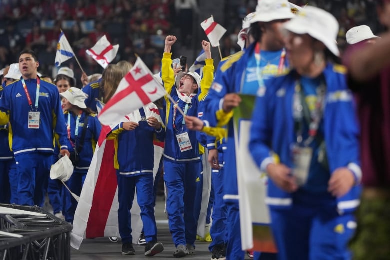 athletes wearing blue and yellow track suits march with red and white flags. A boy in the centre has his arms raised and his mouth wide open/