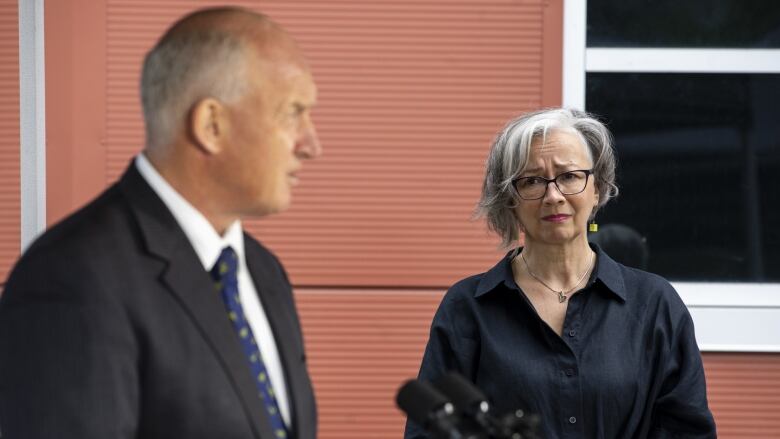 A white man speaks at a podium, with a white woman in the background looking sad.