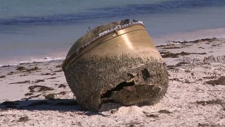 A bronze cylindrical object is shown at the edge of the water on the sand.