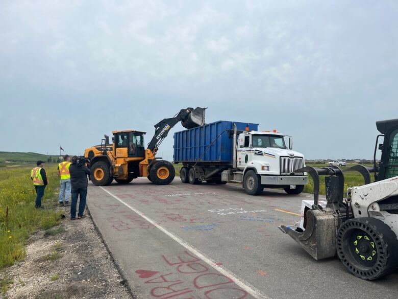 A backhoe shovels something into the back of a truck as people in fluorescent vests watch.